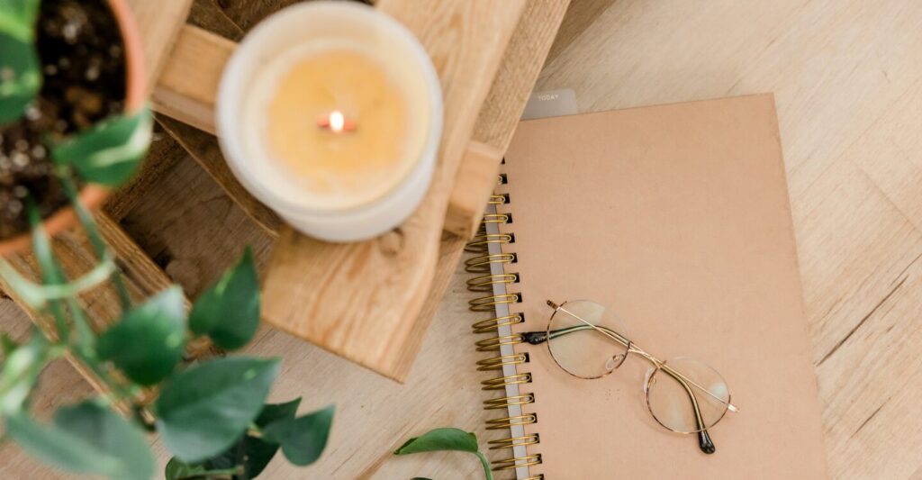 A cozy scene with a lit candle, a spiral notebook, and glasses on a wooden surface.