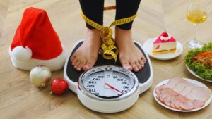 Festive image of bare feet on a bathroom scale surrounded by holiday foods (cake, meat, salad, wine), a Santa hat, ornaments, and a measuring tape around the ankles, symbolizing holiday weight concerns from Sandy Zeldes.