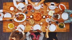 Diverse family gathered around a Christmas table from above view, sharing holiday meal with turkey, pumpkin pie, vegetables, and phones taking photos. Focus: holiday overeating and social feasting. Sandy Zeldes branding.
