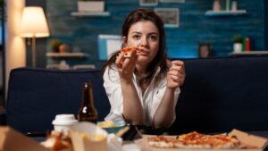 Woman eating pizza on couch at night with snacks and drinks; EFT root-cause approach to binge eating when diets fail.