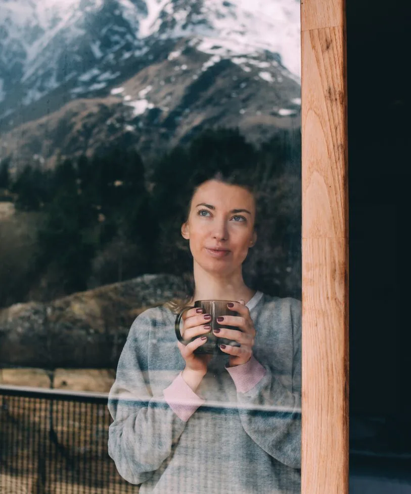 A woman gazing through a window while holding a cup, with a mountain landscape in the background.