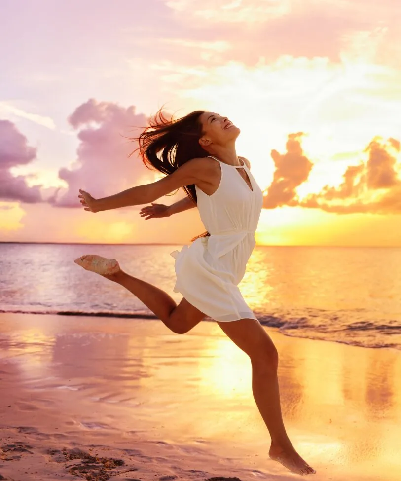 A joyful woman in a white dress running along the beach at sunset with arms outstretched.